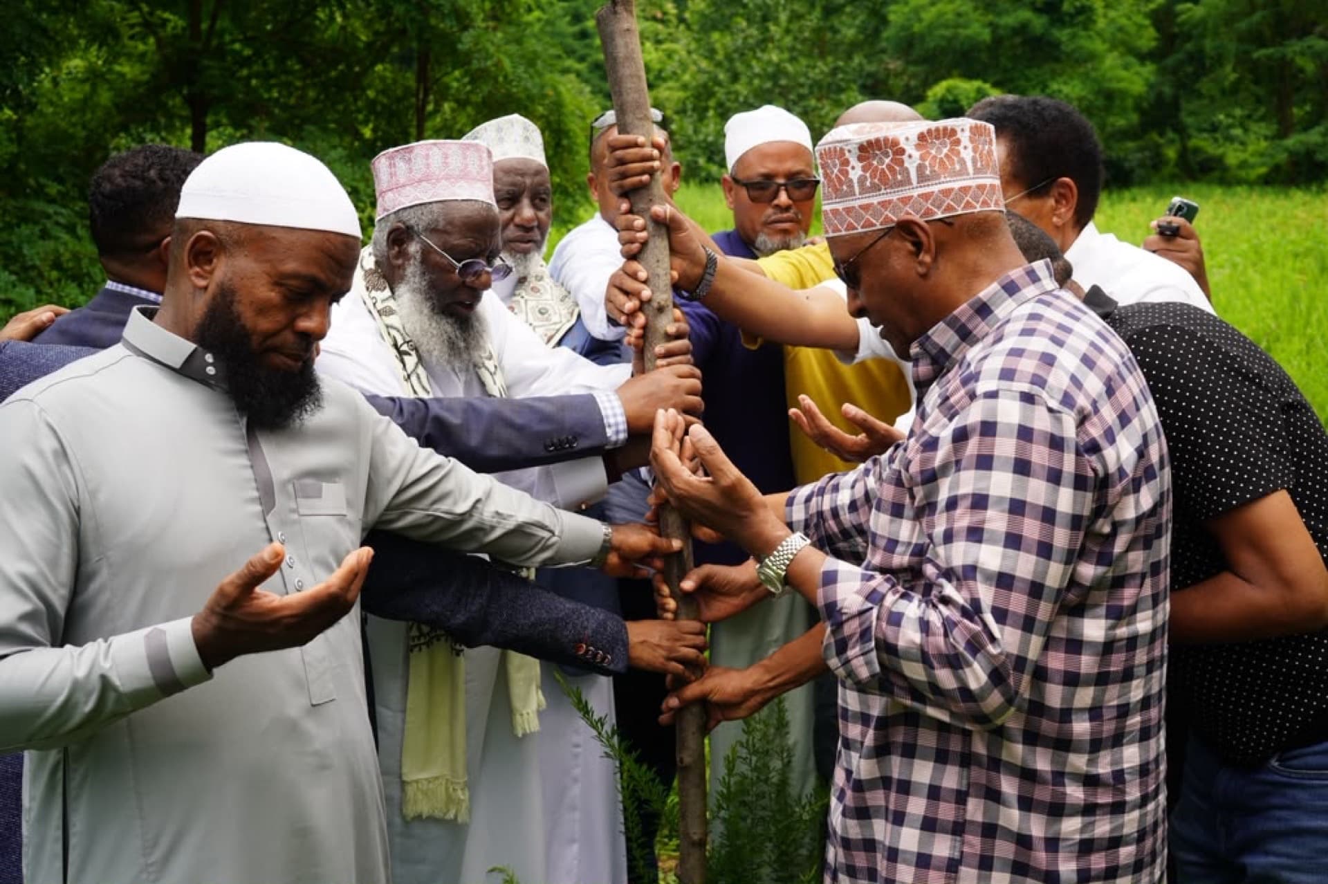 Community elders gather outdoors in dua, hands joined around a planted wooden stake